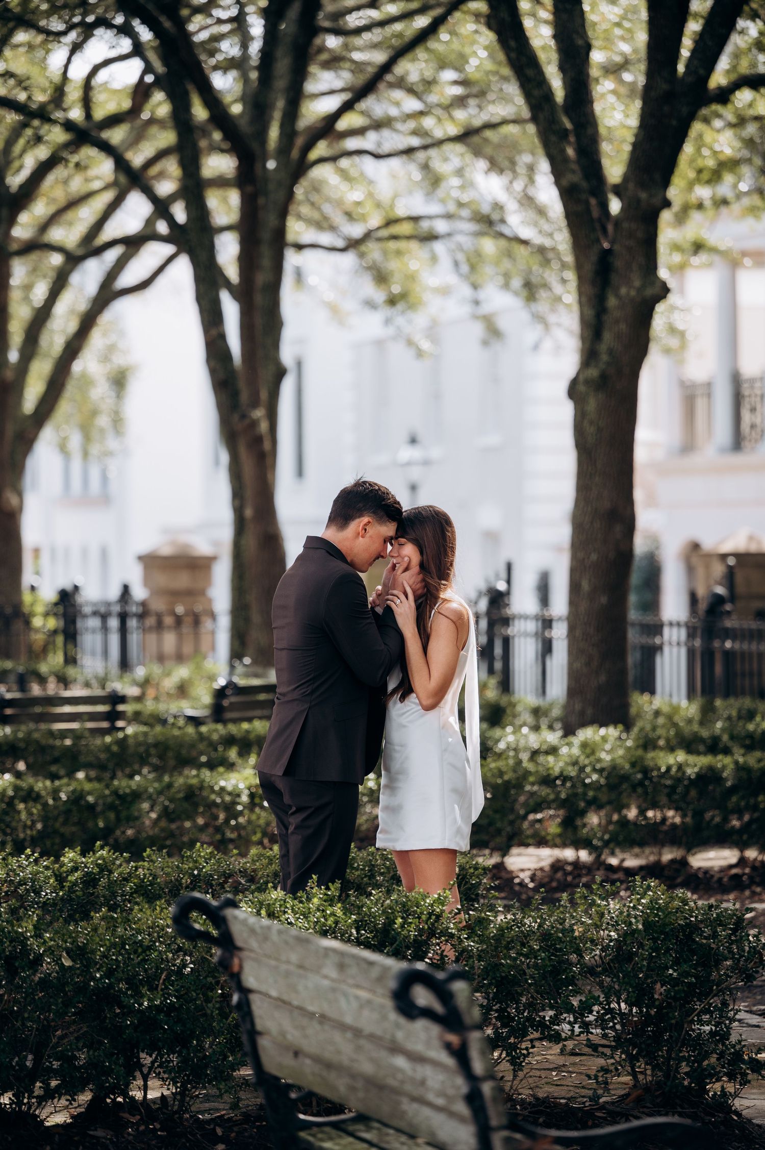 A romantic moment captured between two people under a canopy of trees in an urban park setting.