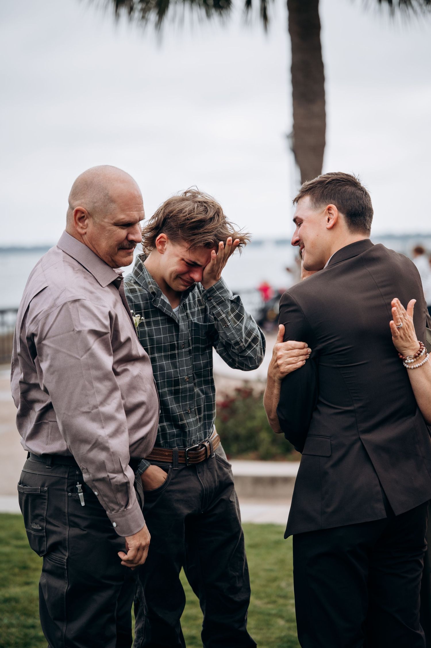 Three people share an emotional conversation outdoors by the waterfront.