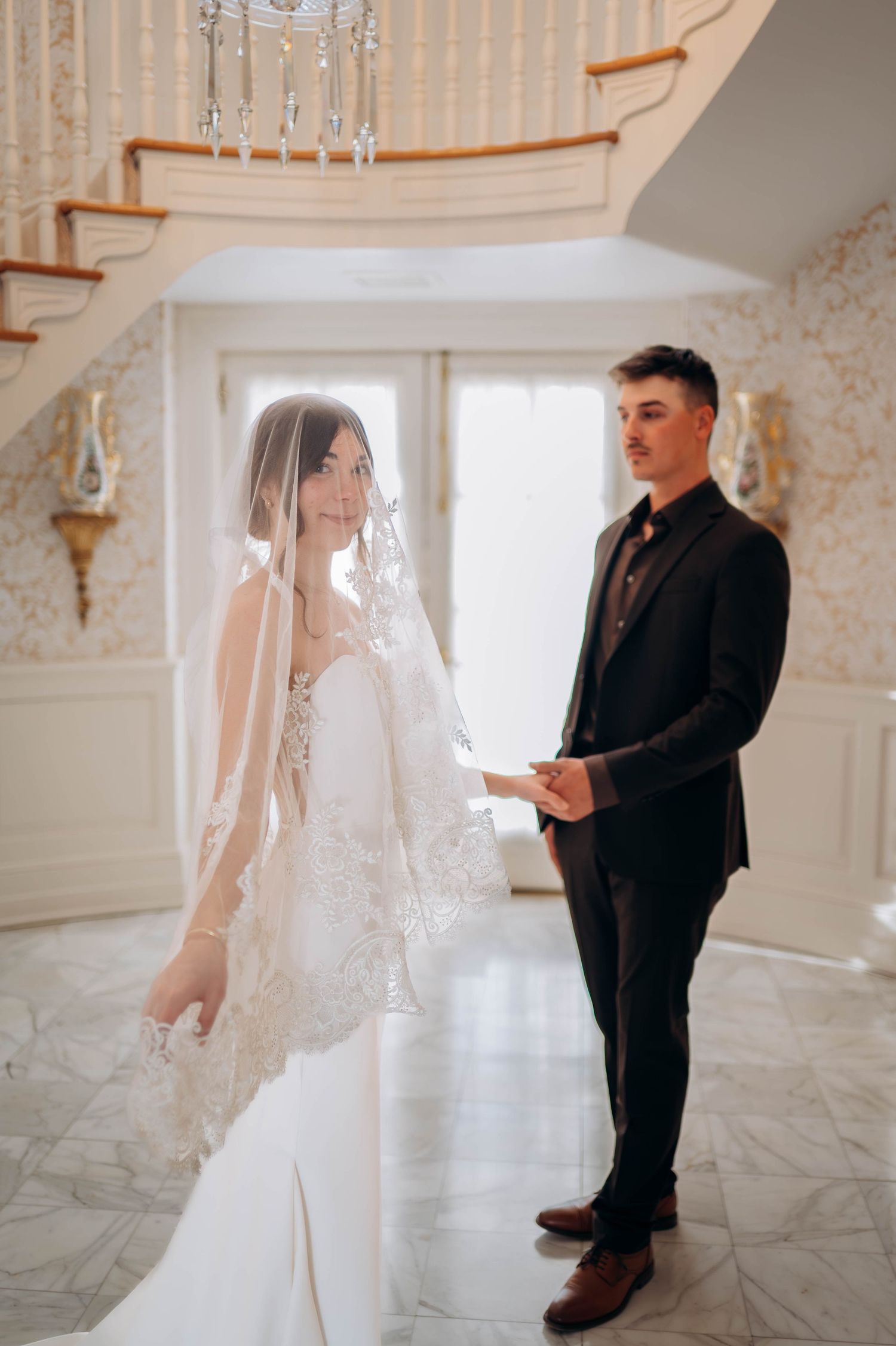 Bride in lace veil and groom exchanging vows in elegant white marble foyer.