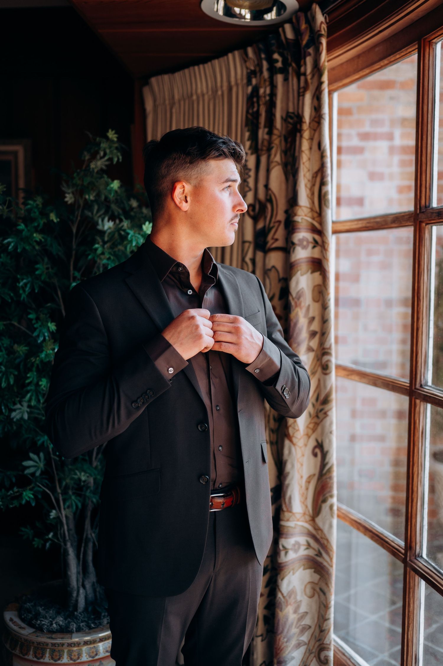 Groom in dark suit adjusting jacket collar while gazing out large window with brick wall backdrop.