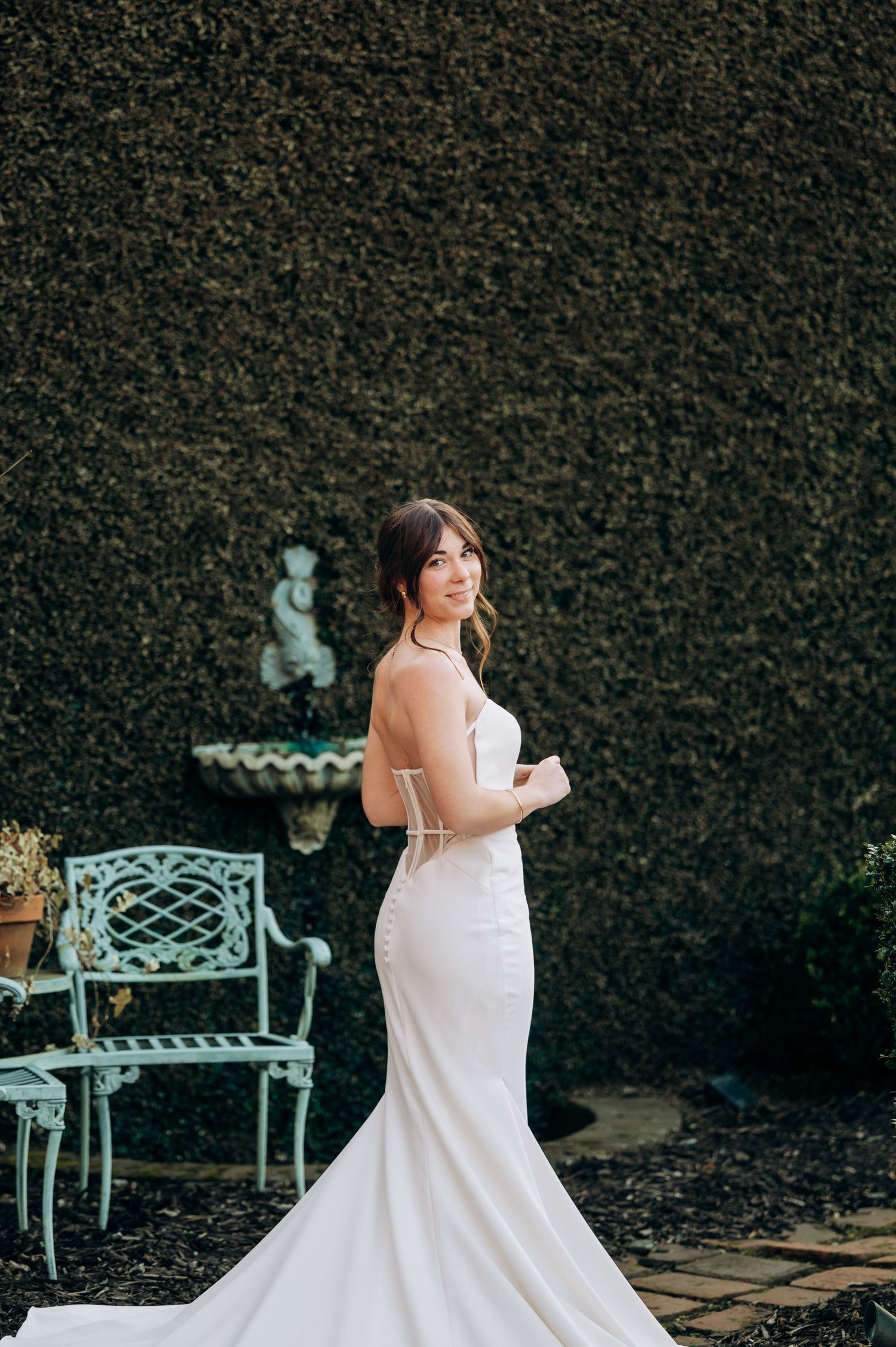 Bride in elegant white mermaid wedding gown posing in garden with ivy wall and vintage teal furniture.