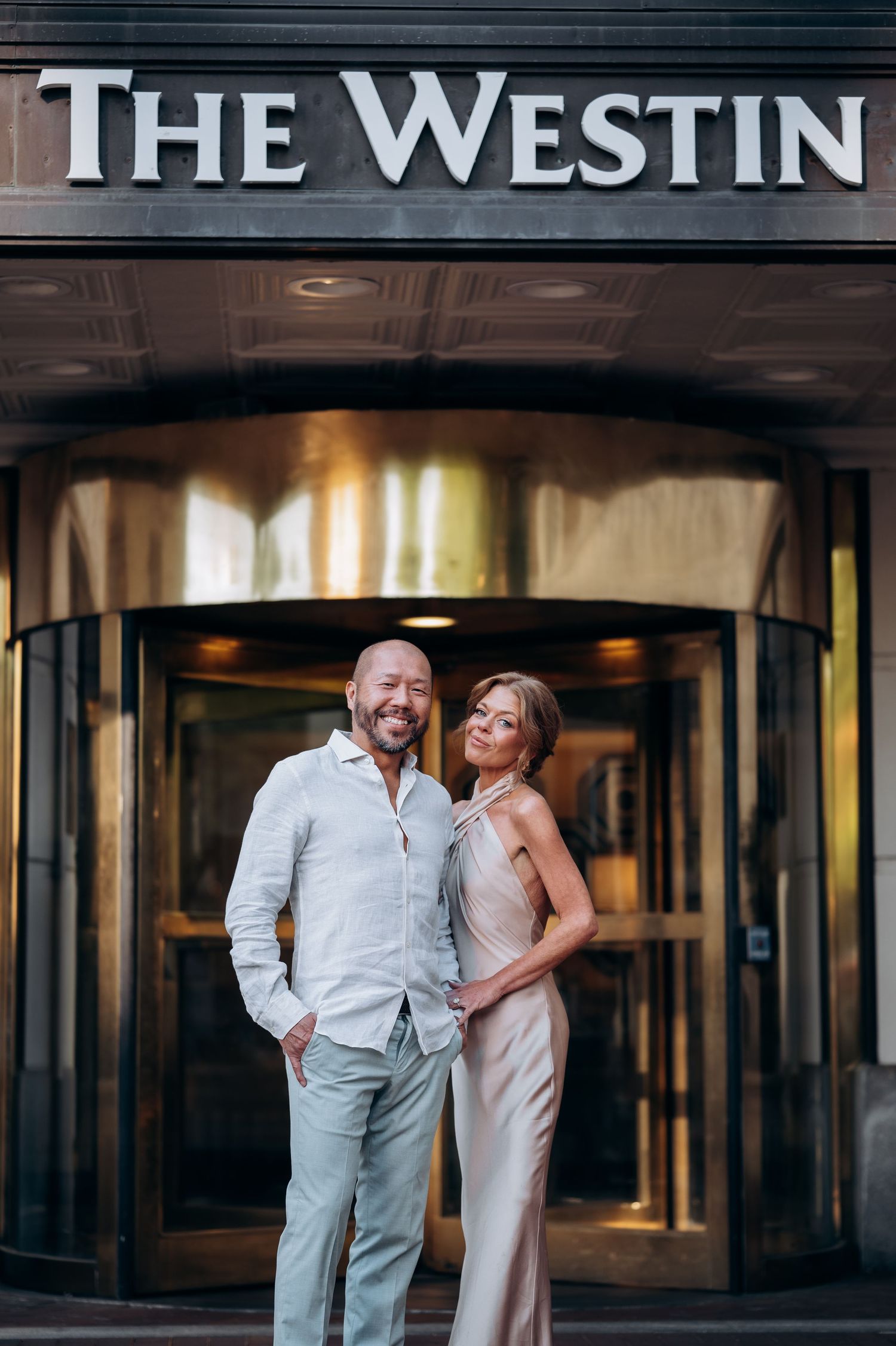 Elegant couple posing in front of The Westin hotel entrance, man in white shirt and woman in satin gown.
