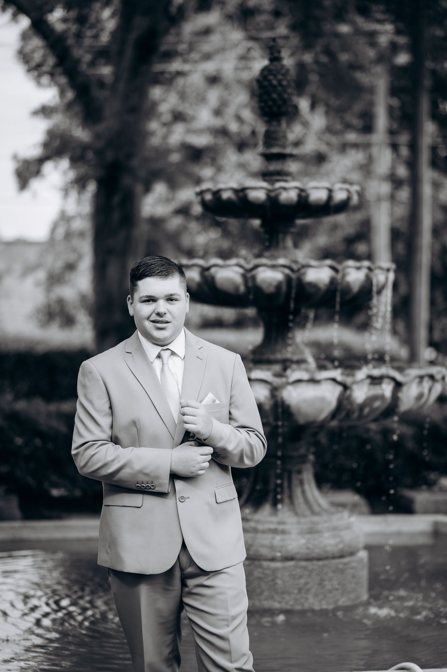 Young man in light gray suit poses confidently by ornate stone fountain in black and white formal portrait.