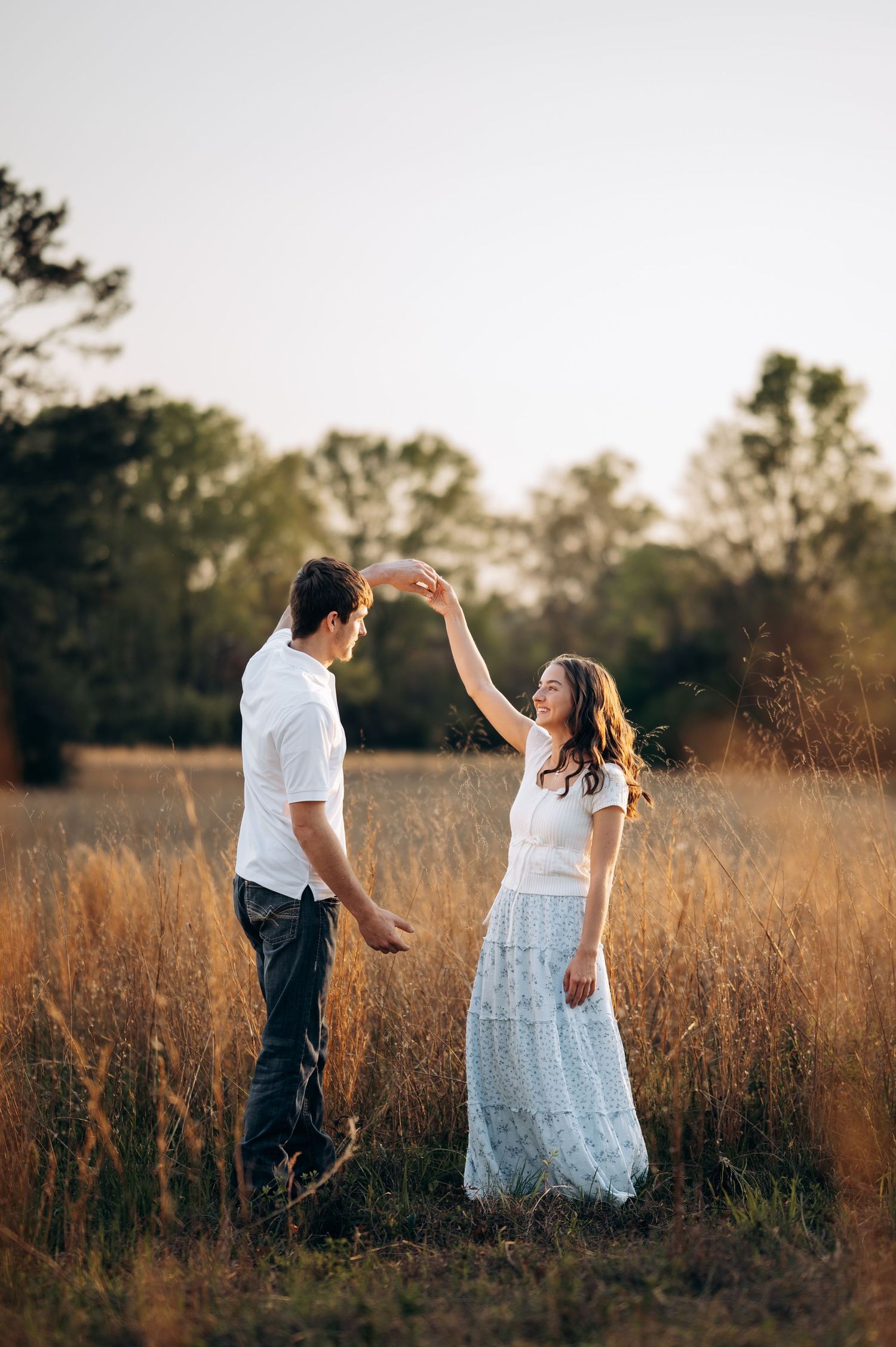A couple dances joyfully in a golden field at sunset, the woman in a white lace skirt twirling gracefully.