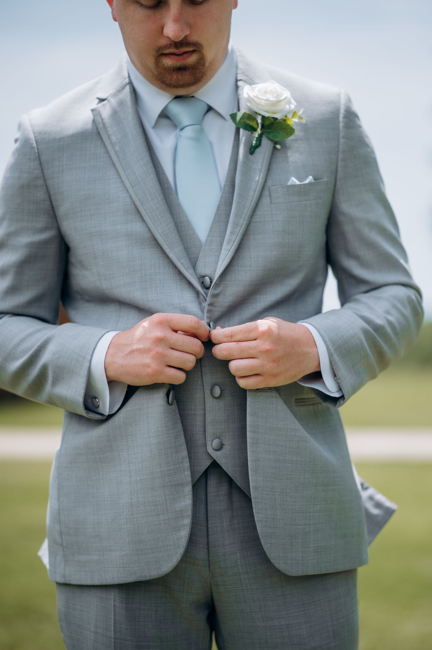 Groom in light gray three-piece suit with mint tie and white rose boutonniere buttoning jacket outdoors.
