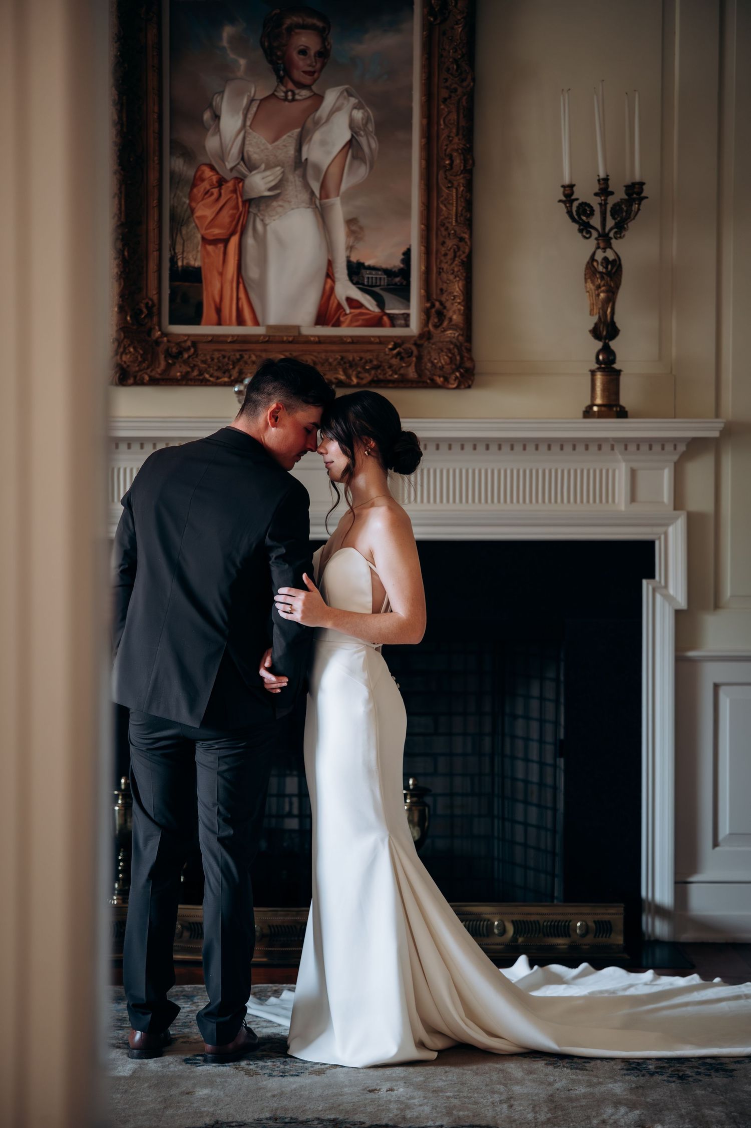 Bride and groom share an intimate moment by a fireplace, with a classic portrait painting above them in an elegant venue.