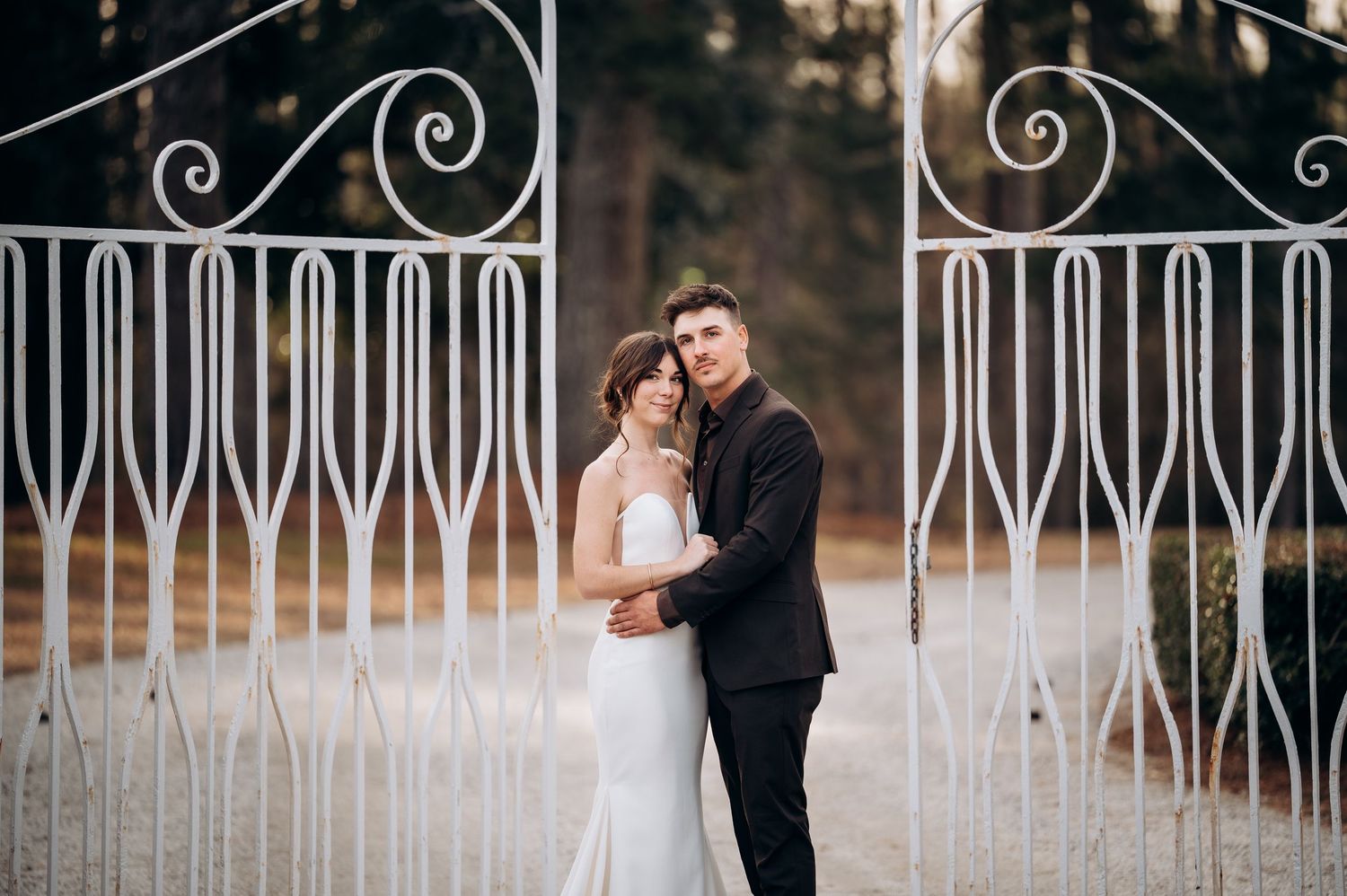 Elegant wedding couple embracing between decorative white iron gates at sunset.