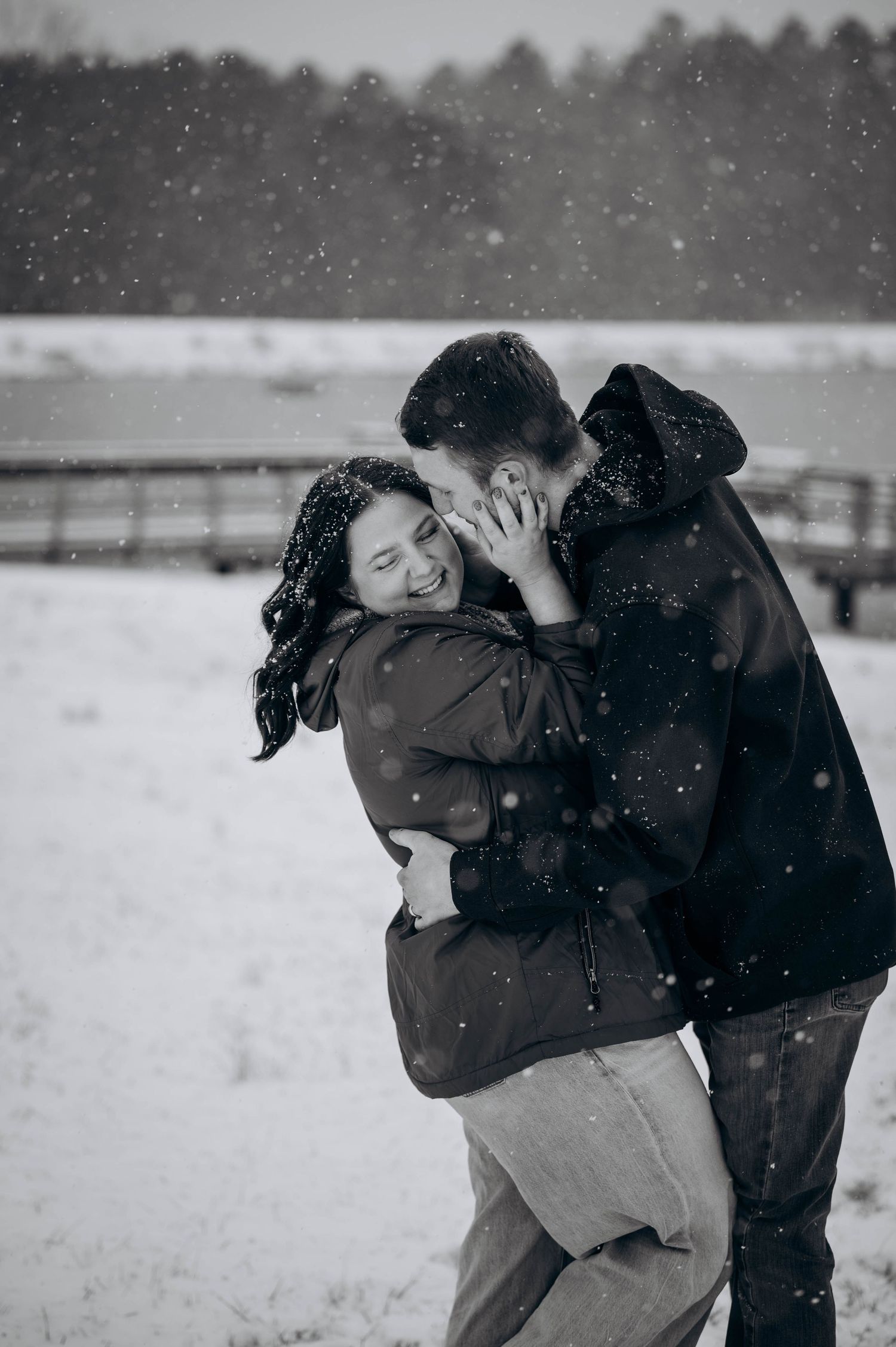 A romantic couple embraces in the falling snow with a winter landscape and fence in the background.