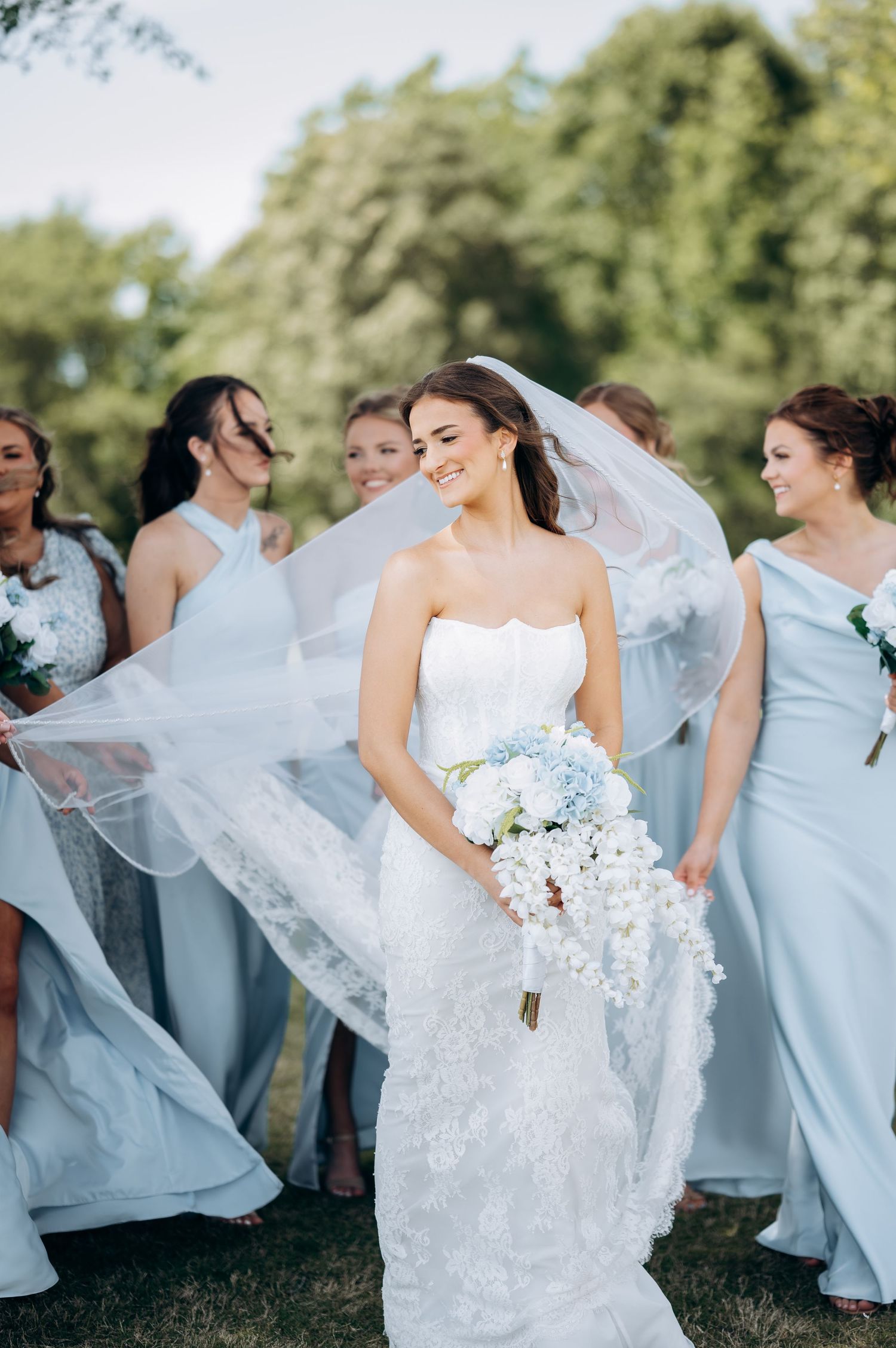 Smiling bride in white lace gown holds blue bouquet as veil flows, surrounded by bridesmaids in light blue dresses outdoors.