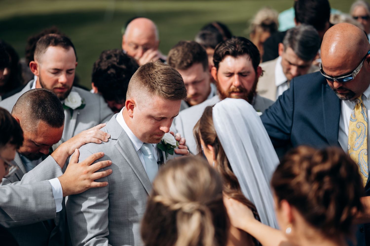 Emotional outdoor wedding prayer circle surrounds bride and groom during heartfelt ceremony moment.