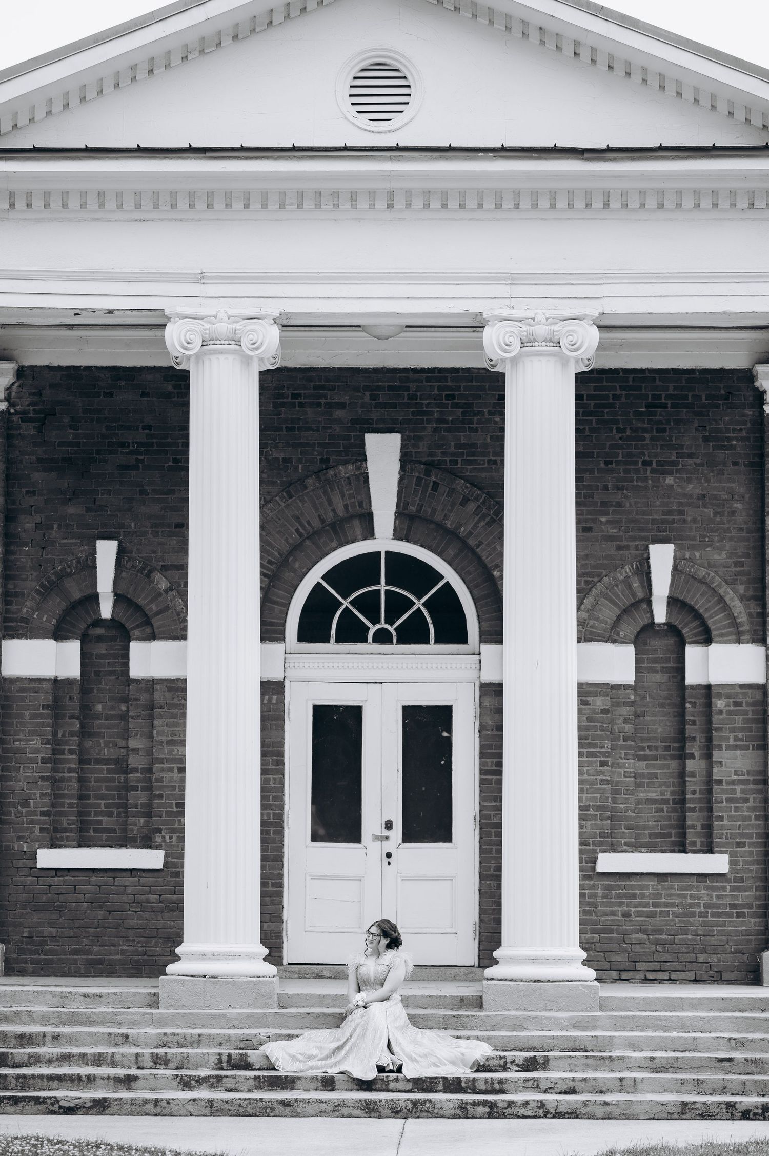 Bride in wedding gown sitting on steps of classic brick building with white columns and arched doorway.