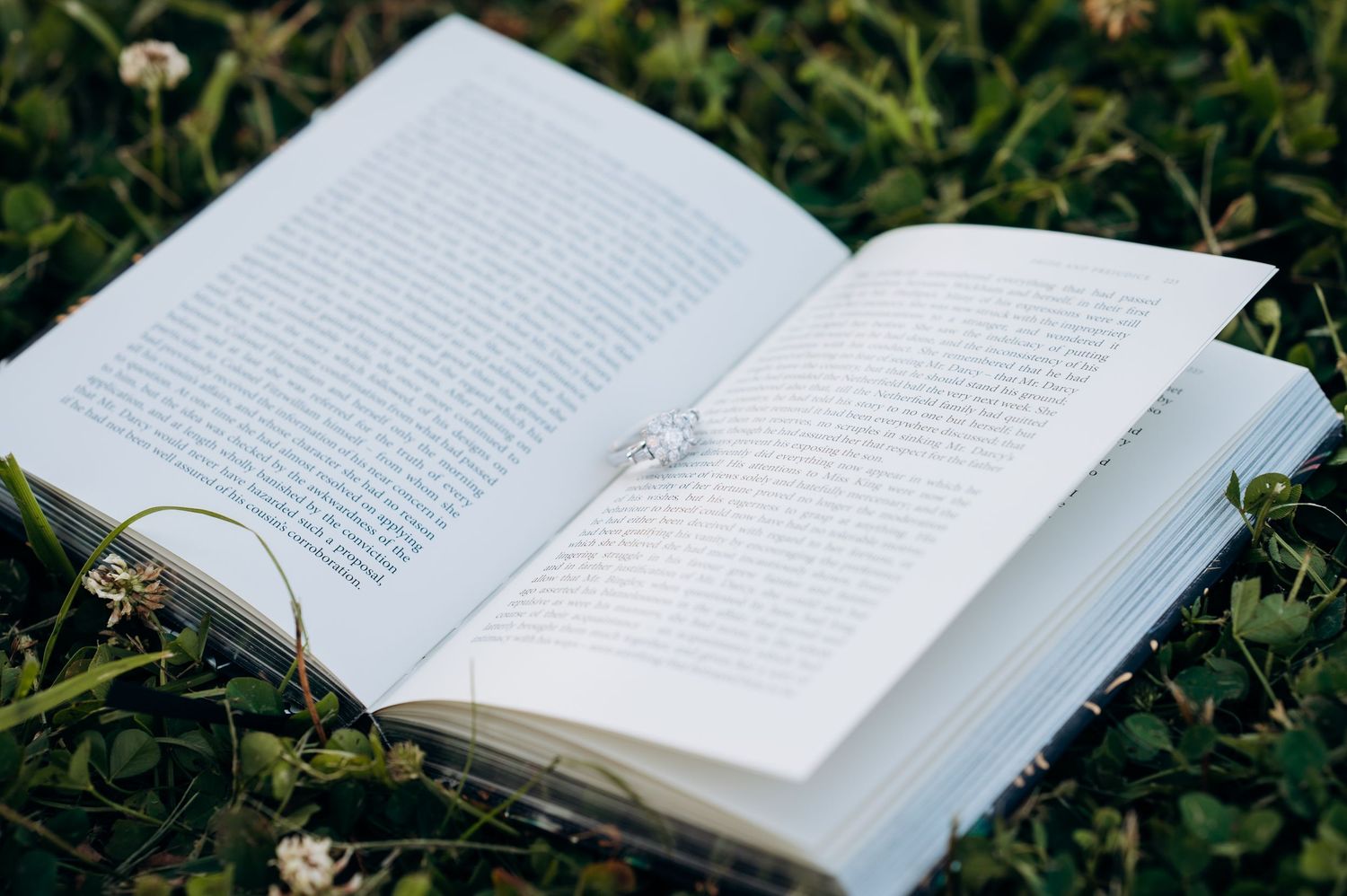 Open book with a diamond ring resting on its pages, lying on lush green grass outdoors.