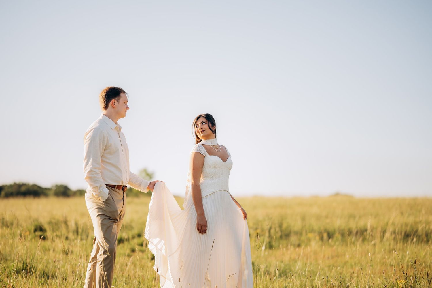 Bride and groom holding hands in a sunlit open field, bride wearing a flowing white gown on their wedding day.