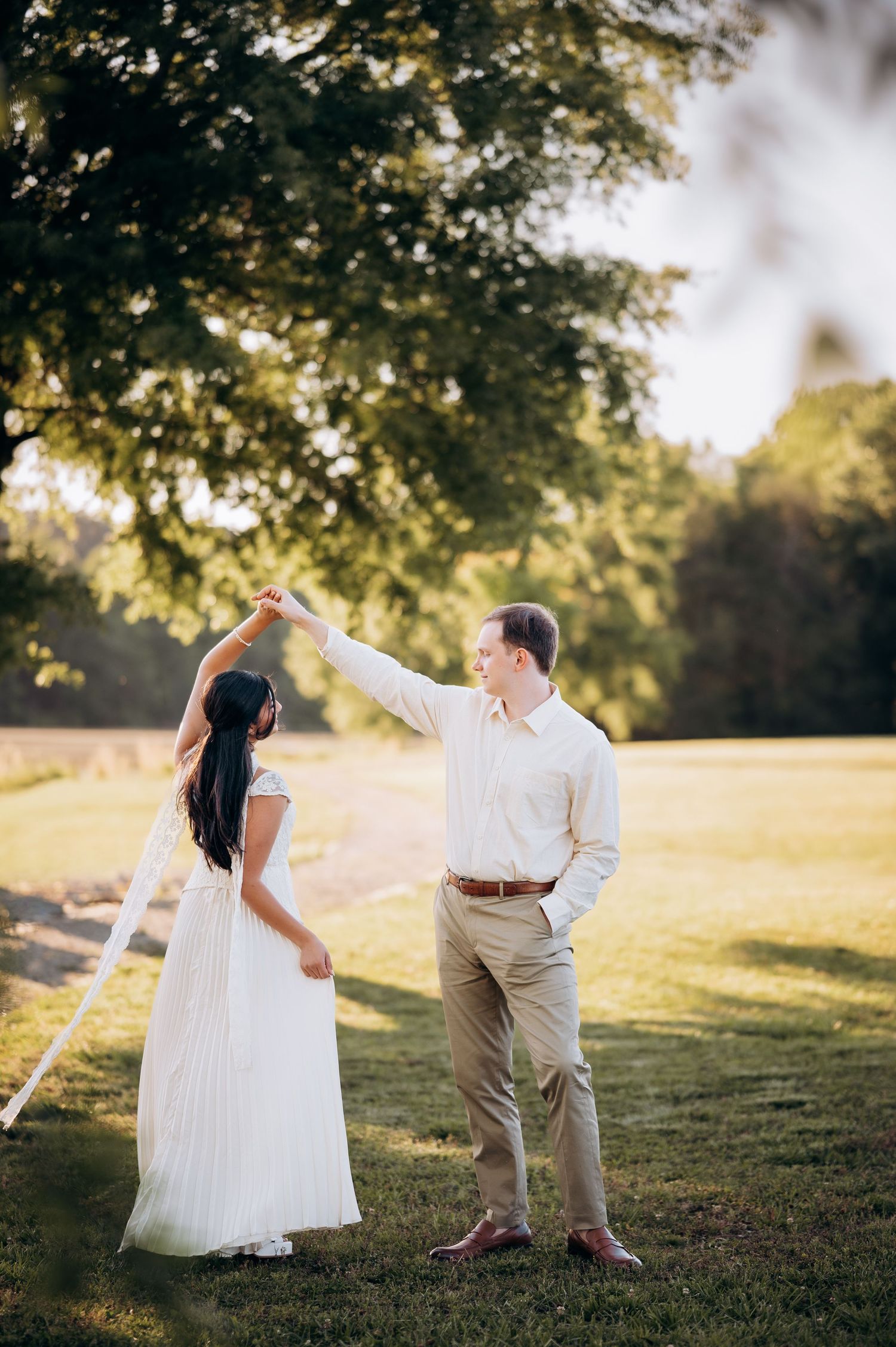 A couple dances outdoors on a sunny lawn, the woman in a white gown being twirled by a man in khaki pants.