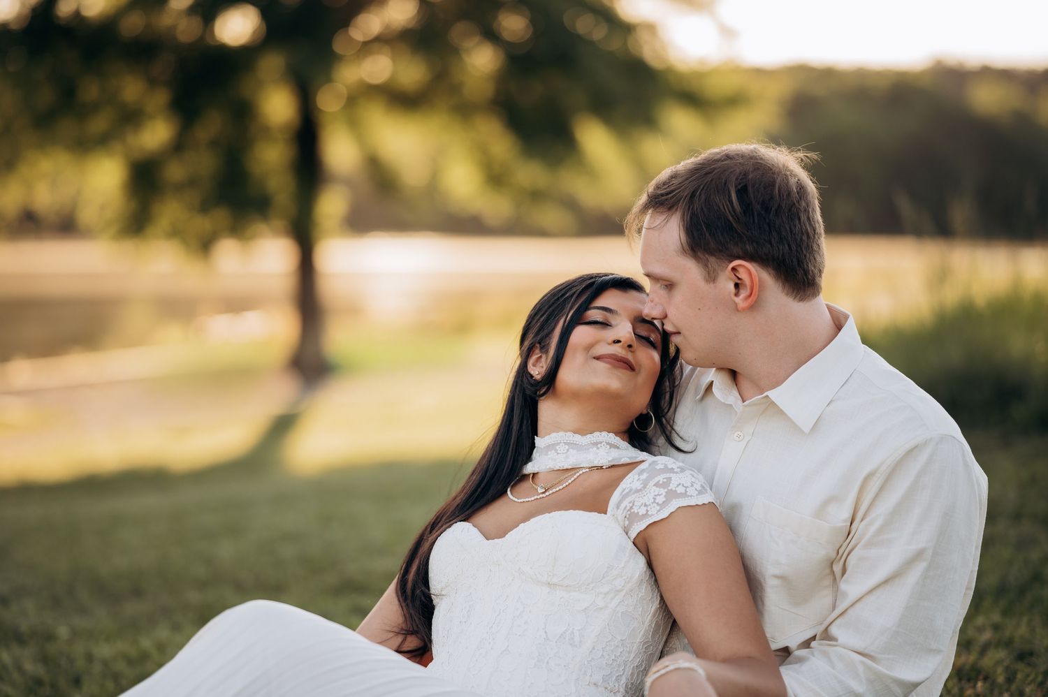Couple sharing a romantic moment outdoors at golden hour, woman in white dress leaning back against man on green grass.
