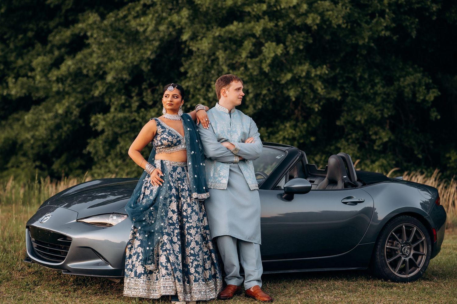 Couple in elegant Indian attire posing by a gray Mazda MX-5 convertible in a lush outdoor setting.