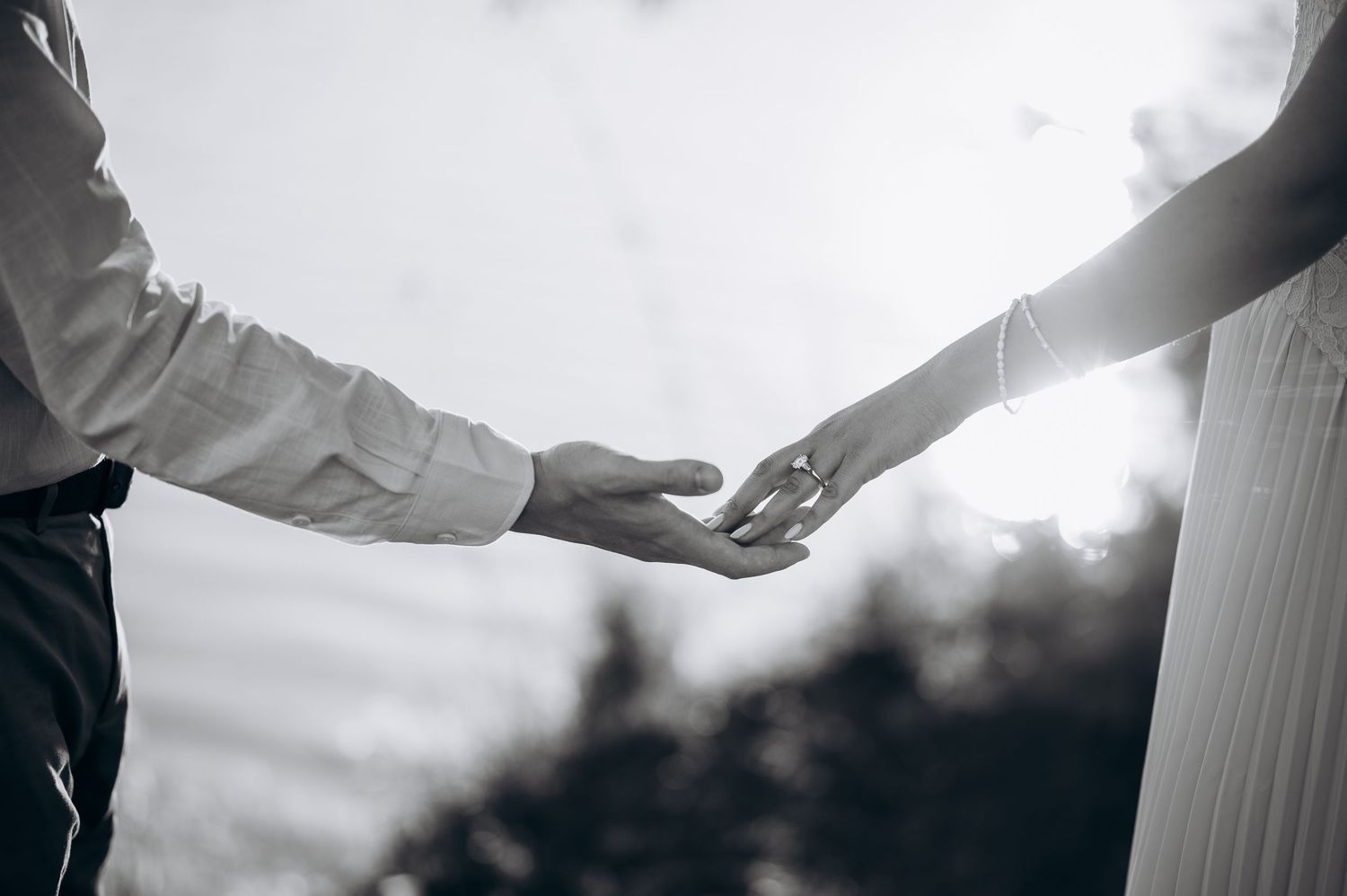 Black and white photo of a couple gently reaching for each other's hands outdoors in soft natural light.