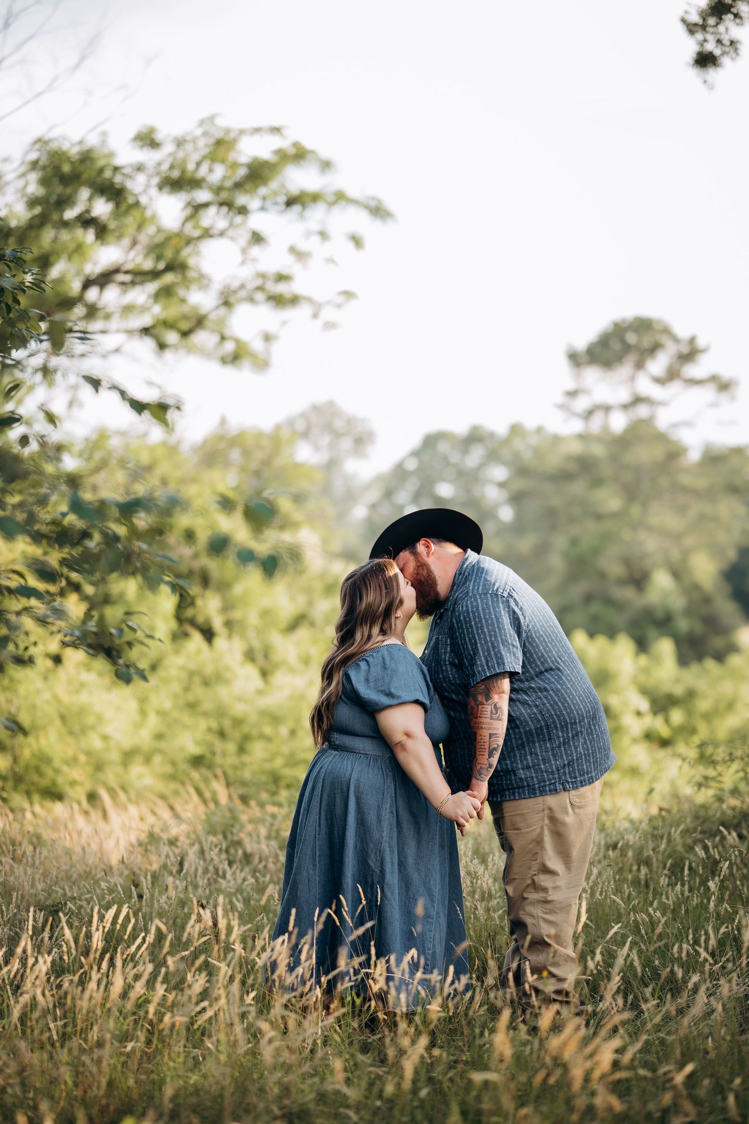 Couple sharing a romantic kiss in a lush green meadow, woman in blue dress holding hands with tattooed man in black hat.