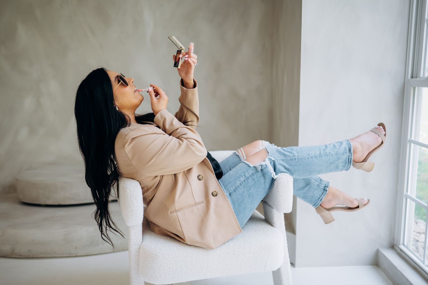 Relaxed person in beige sweater and jeans lounging on white couch by window while applying lip product.