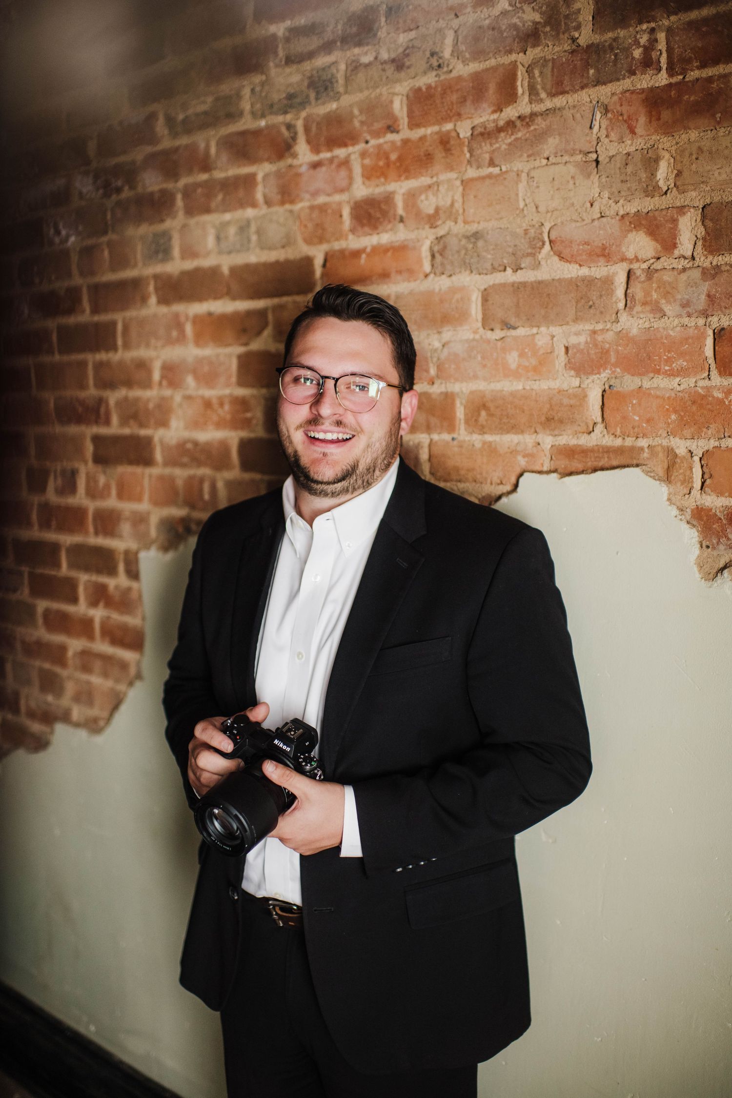 Photographer in formal attire poses with camera against exposed brick wall.