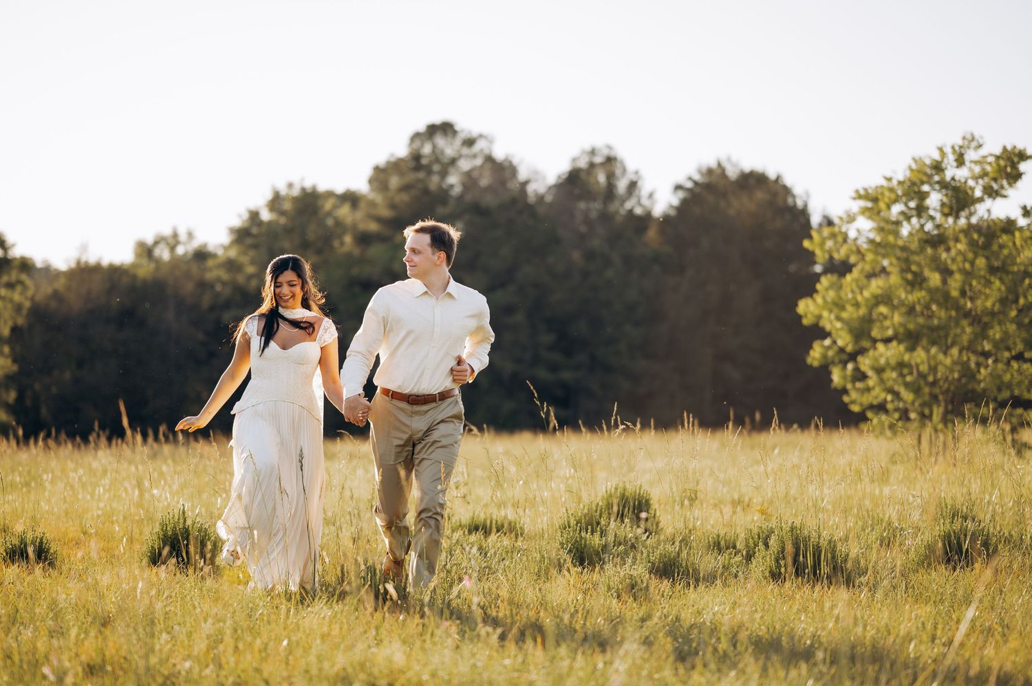 Happy couple strolling hand in hand across a sunny open field surrounded by tall green trees on their wedding day.
