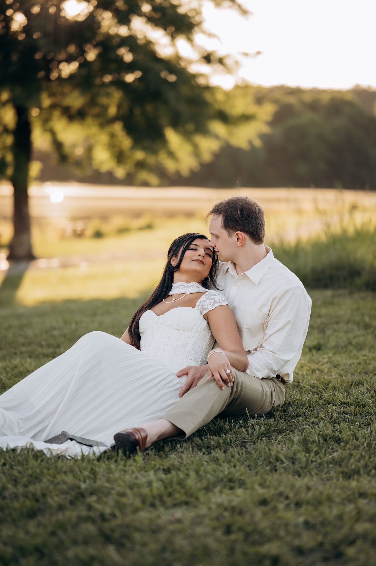 Couple sitting on green grass at sunset, woman in white dress leaning back against man in beige shirt, sharing a romantic moment.