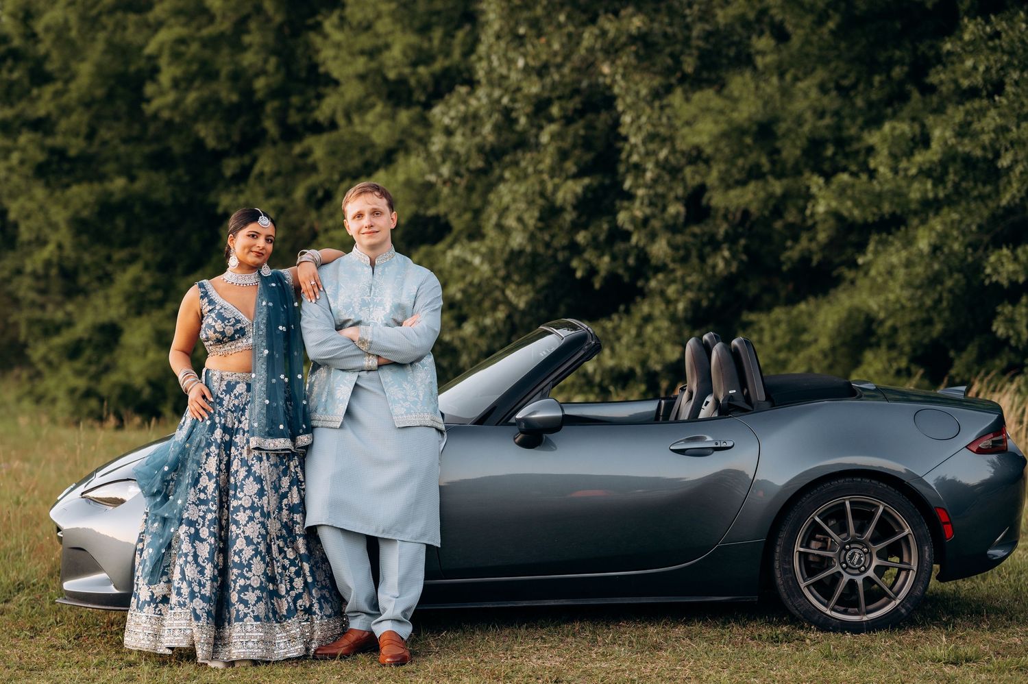 Couple in blue Indian attire poses by a gray Mazda MX-5 Miata convertible with lush green trees behind.