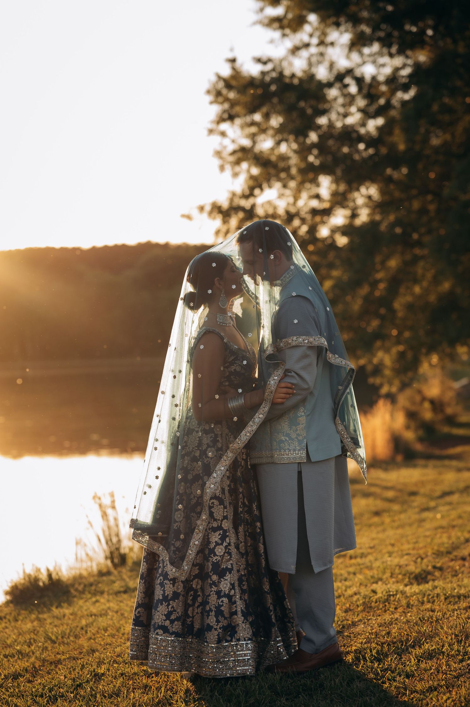 Couple in traditional South Asian wedding attire sharing a romantic moment under a veil at golden hour by a lake.