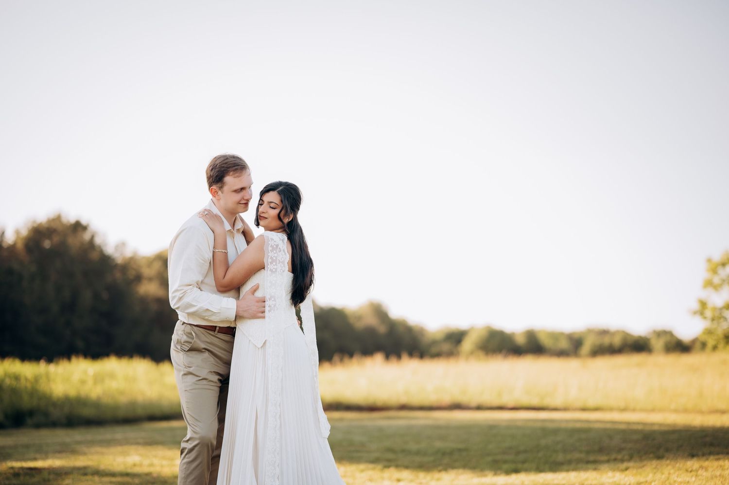 Couple embracing in a sunny field, bride in white gown and groom in tan suit, sharing a romantic outdoor wedding moment.
