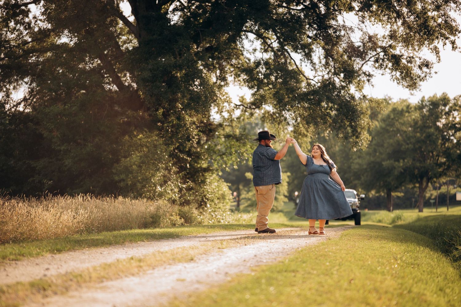Couple dancing on a scenic dirt path surrounded by lush green trees in warm golden sunlight.