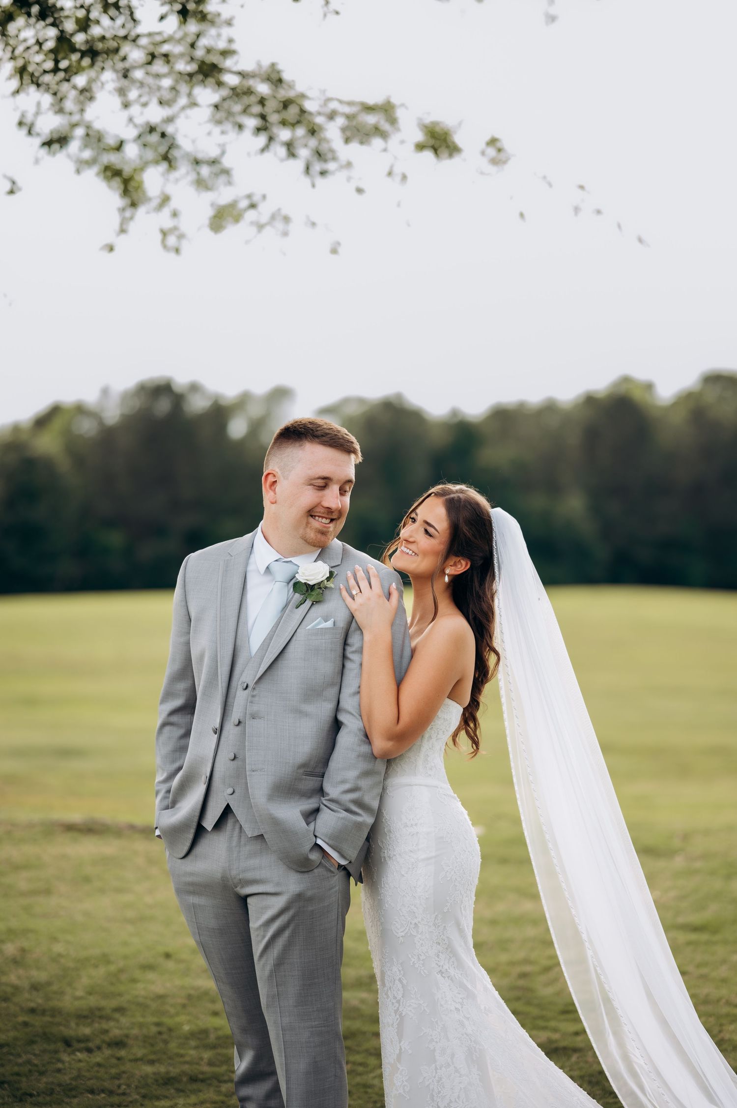 Smiling bride in lace gown with veil leans on groom in gray suit on lush green wedding lawn.