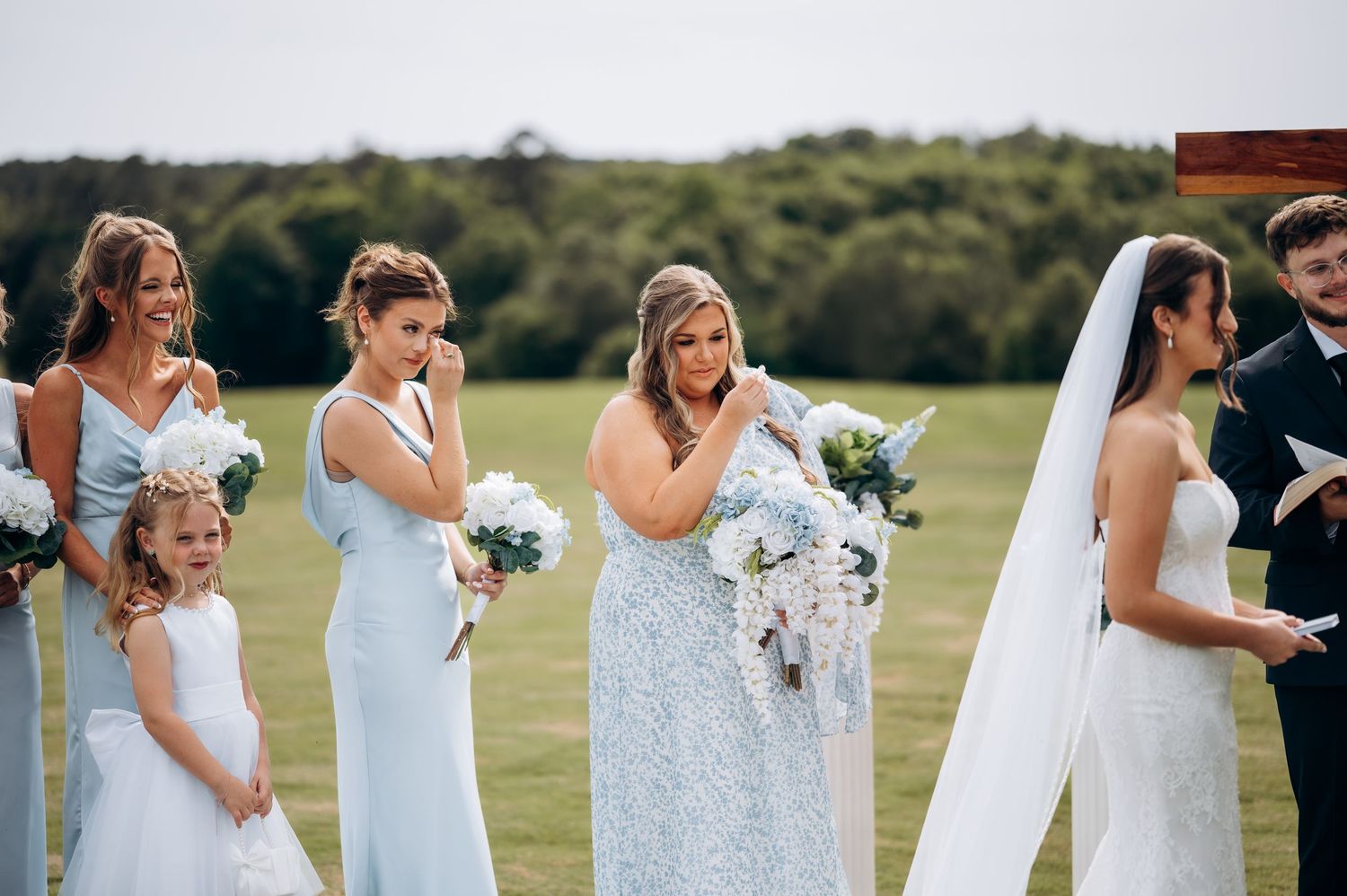 Bridesmaids in light blue dresses crying emotional tears at outdoor wedding ceremony.