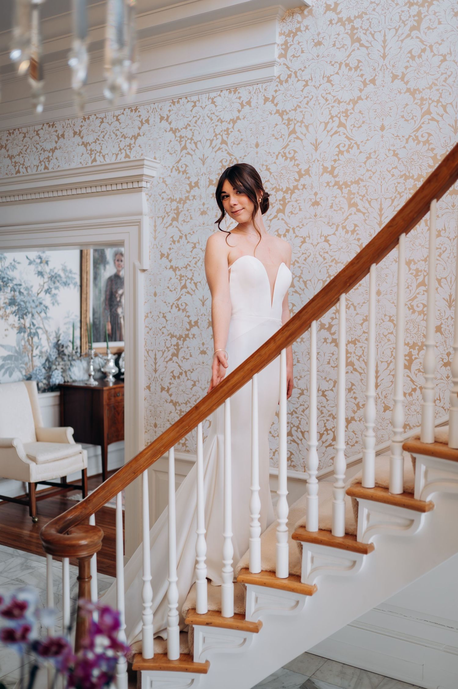 Bride in white gown standing on elegant staircase with ornate wallpaper.