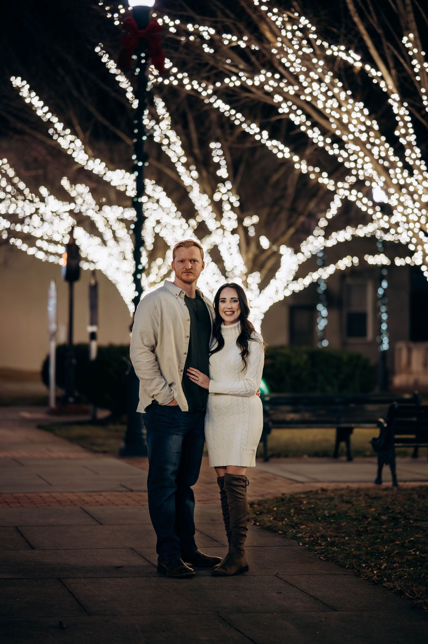 Pregnant couple posing before stunning white light-wrapped trees at night, woman in white sweater dress and tall boots.