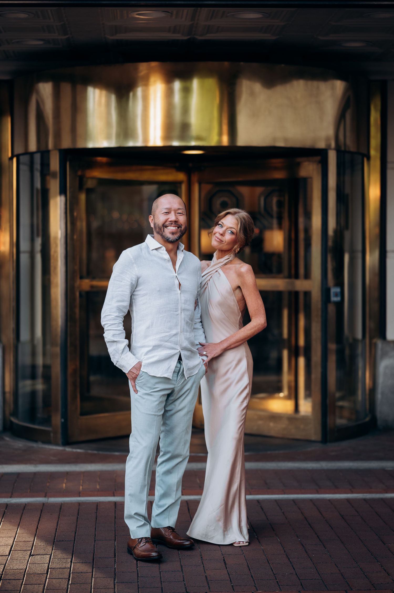 Smiling couple posing in front of a revolving door, man in linen suit and woman in sleek satin dress.