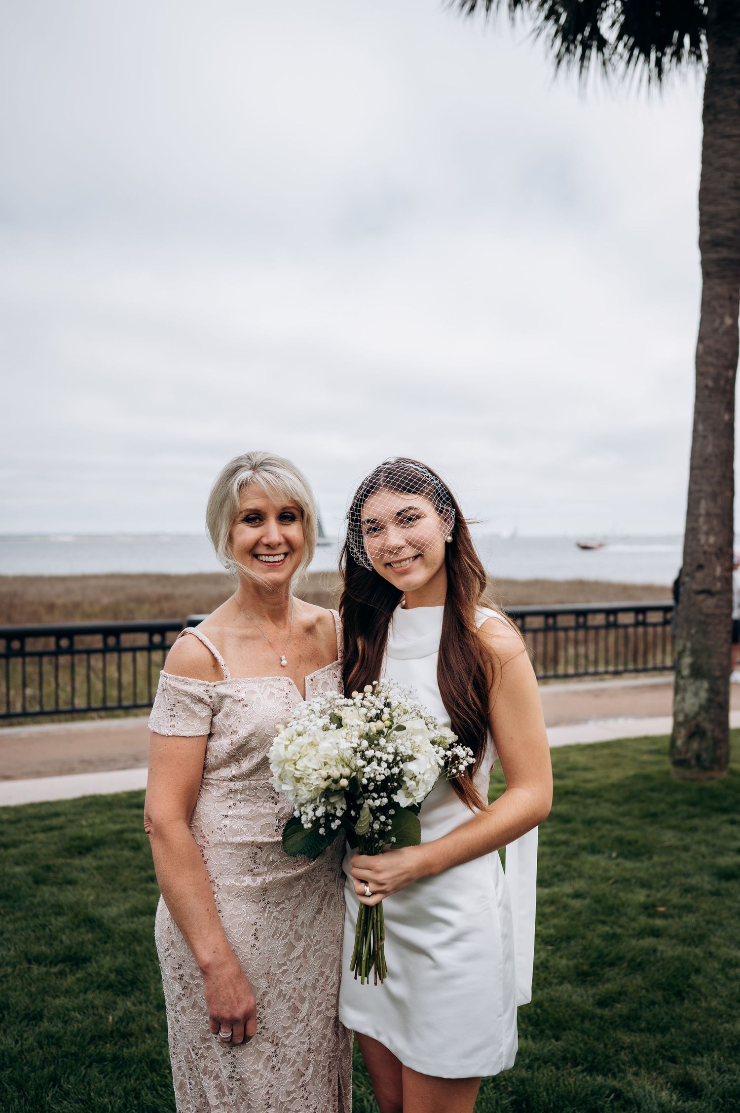 Bride in white mini dress and birdcage veil poses with bouquet alongside older woman in lace dress outdoors.