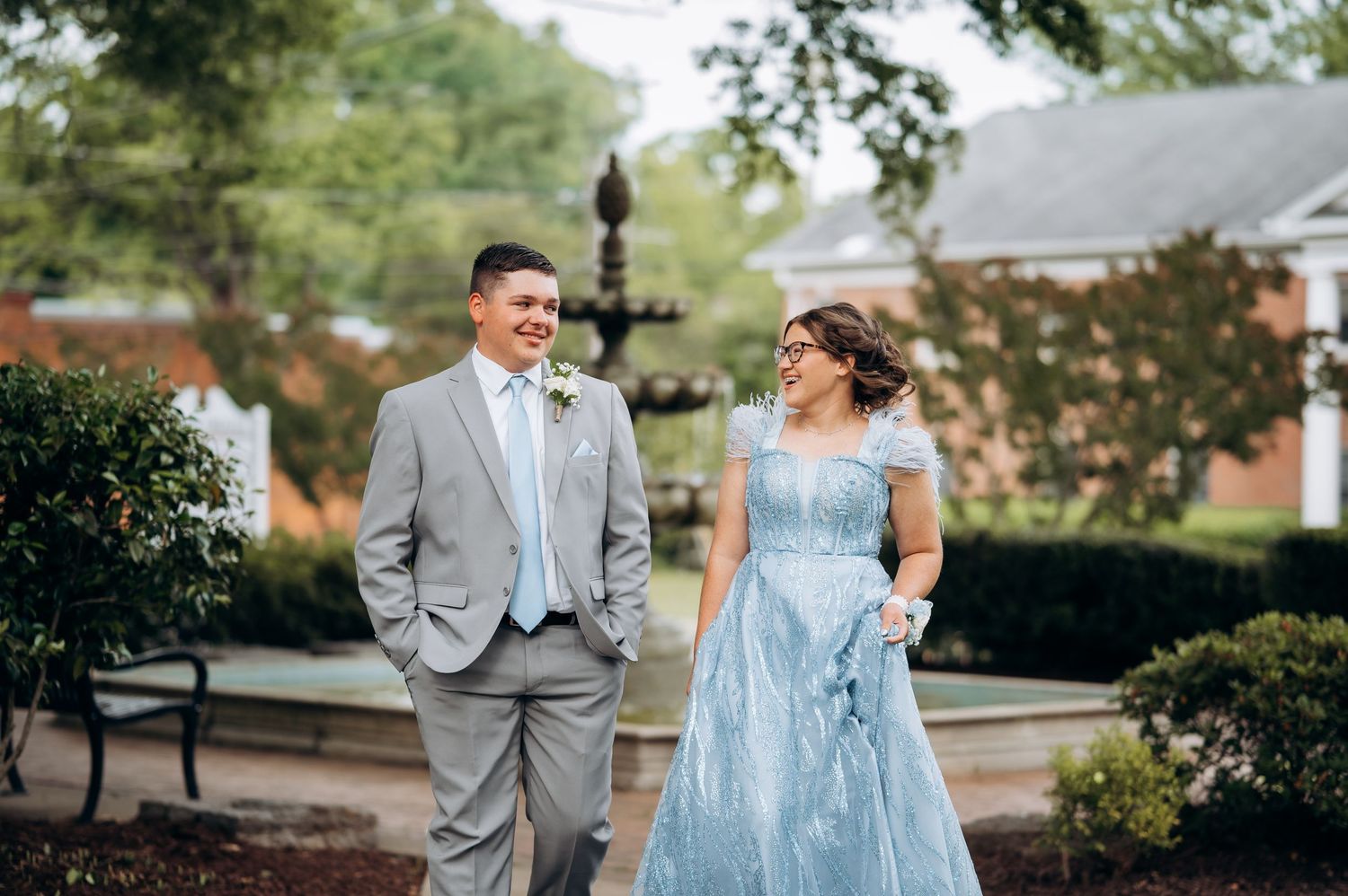 Couple in formal attire, man in gray suit and woman in blue gown, walking outdoors near a fountain.