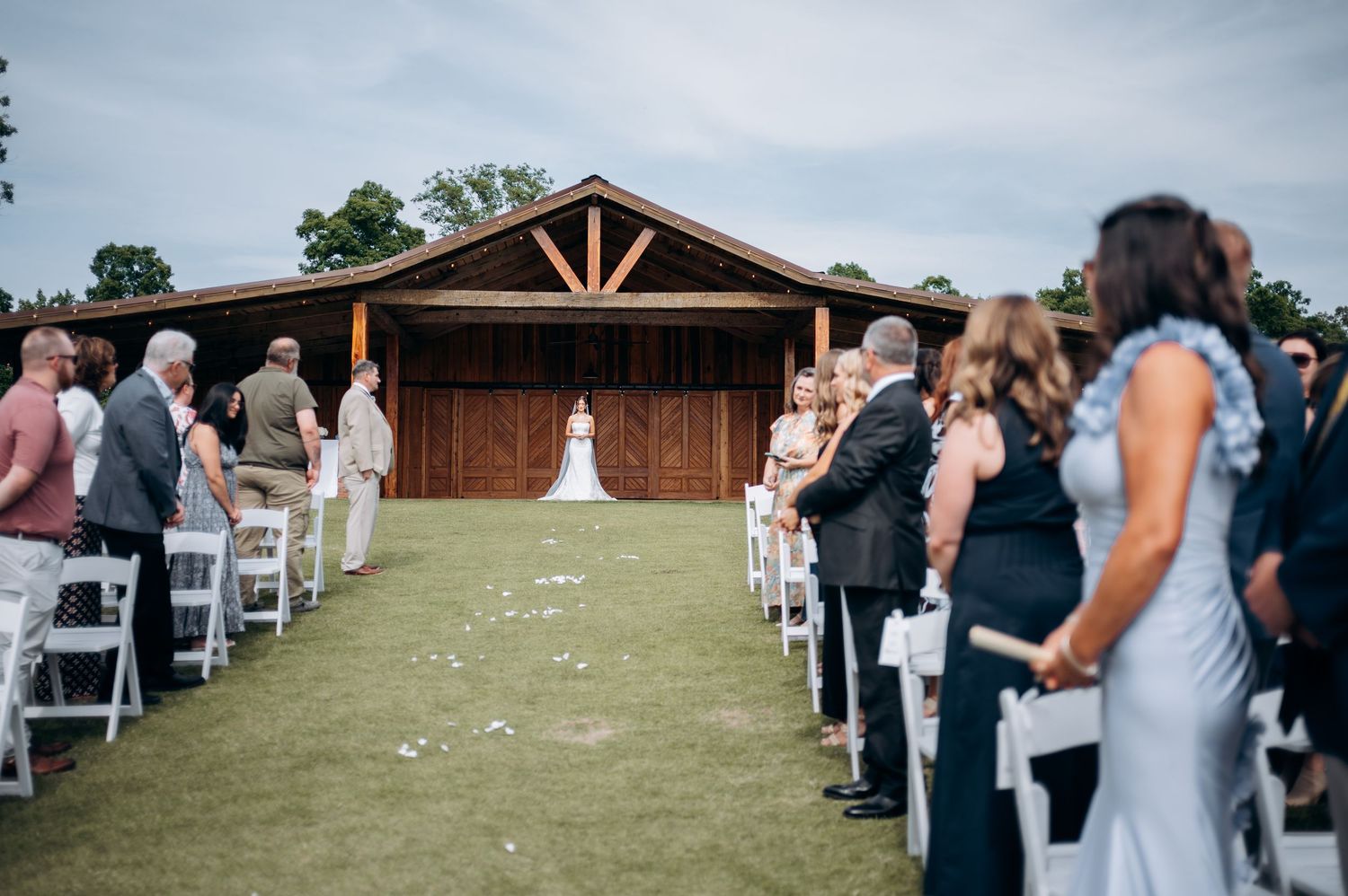 Guests stand as bride walks down the aisle toward a rustic wooden barn at an outdoor wedding.