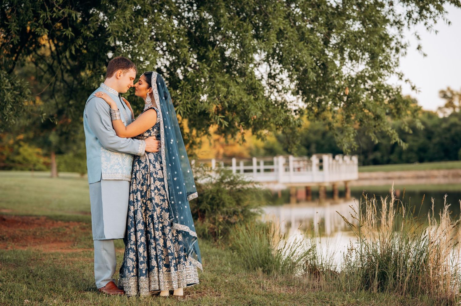 Couple embracing in traditional Indian wedding attire near a serene pond surrounded by lush greenery at golden hour.