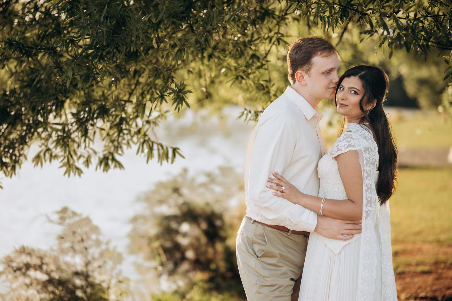 Couple embracing outdoors near a lake under green willow trees, bride in white lace dress, groom in khaki pants.