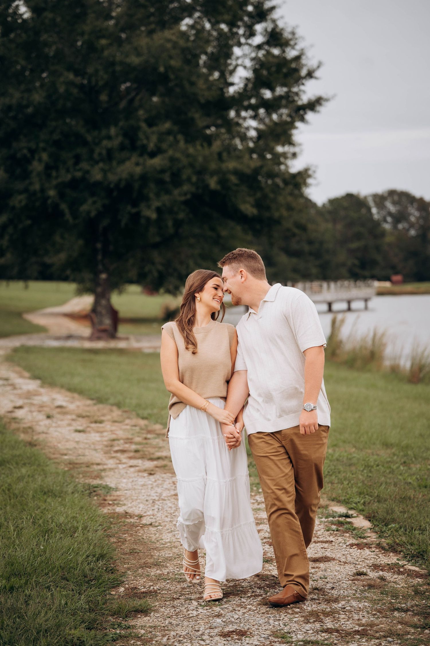 Couple in white and khaki stroll along a lakeside path during an engagement photoshoot.