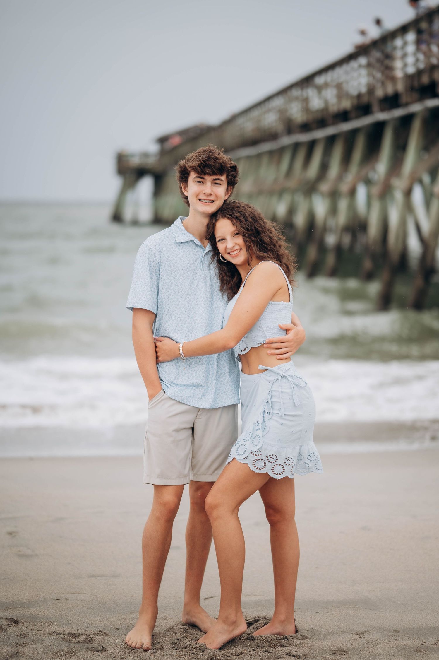 Couple poses together on beach with fishing pier in background.