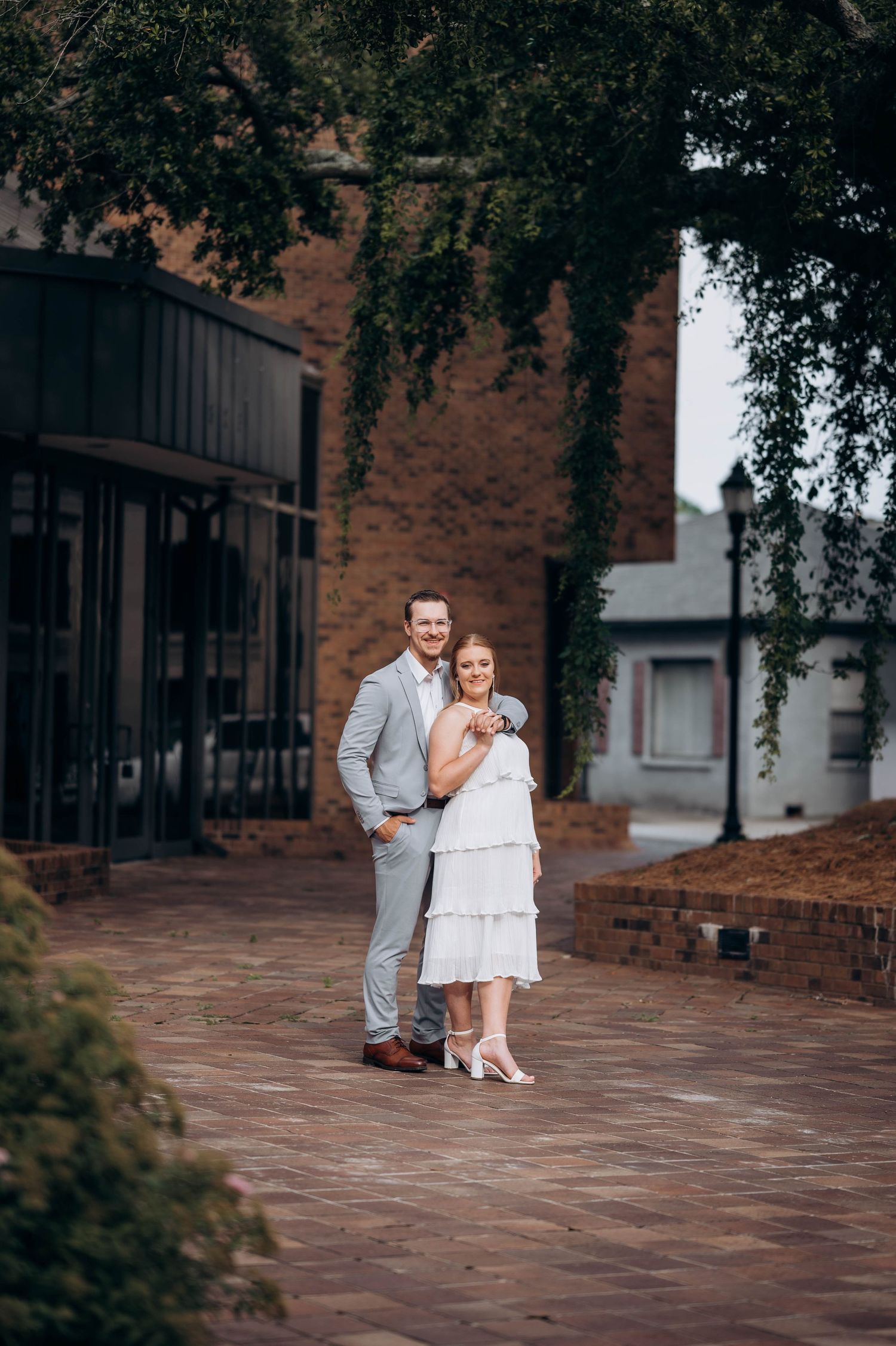A couple poses together on a brick patio near historic buildings.