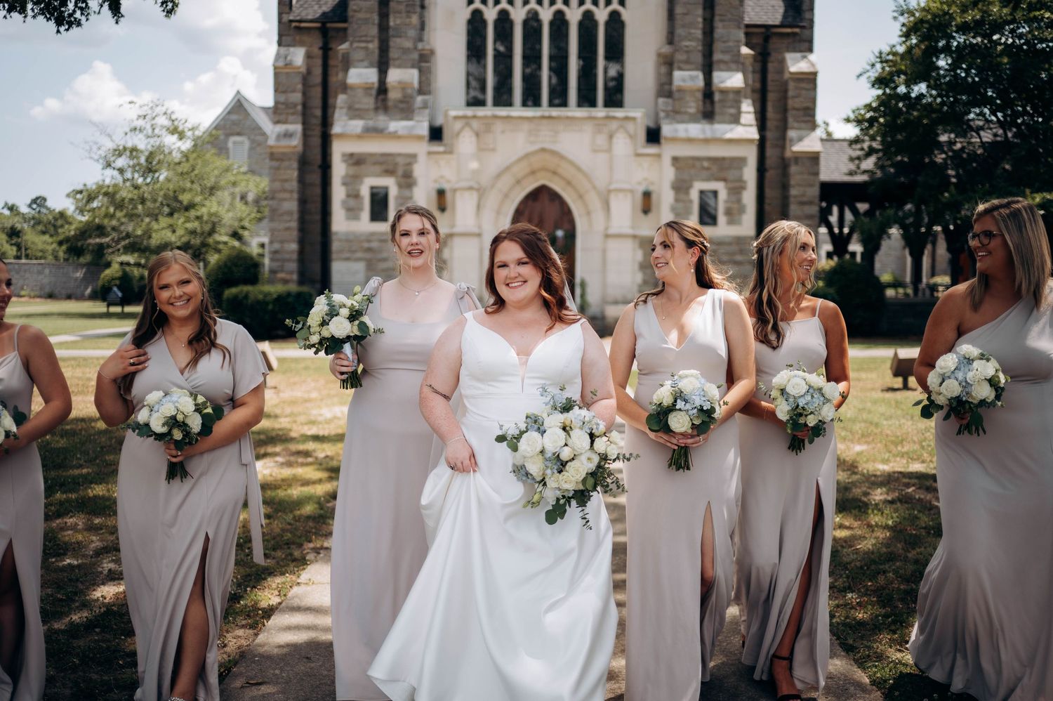 Bridesmaids in light gray dresses stand with bouquets outside a stone church on a sunny wedding day.