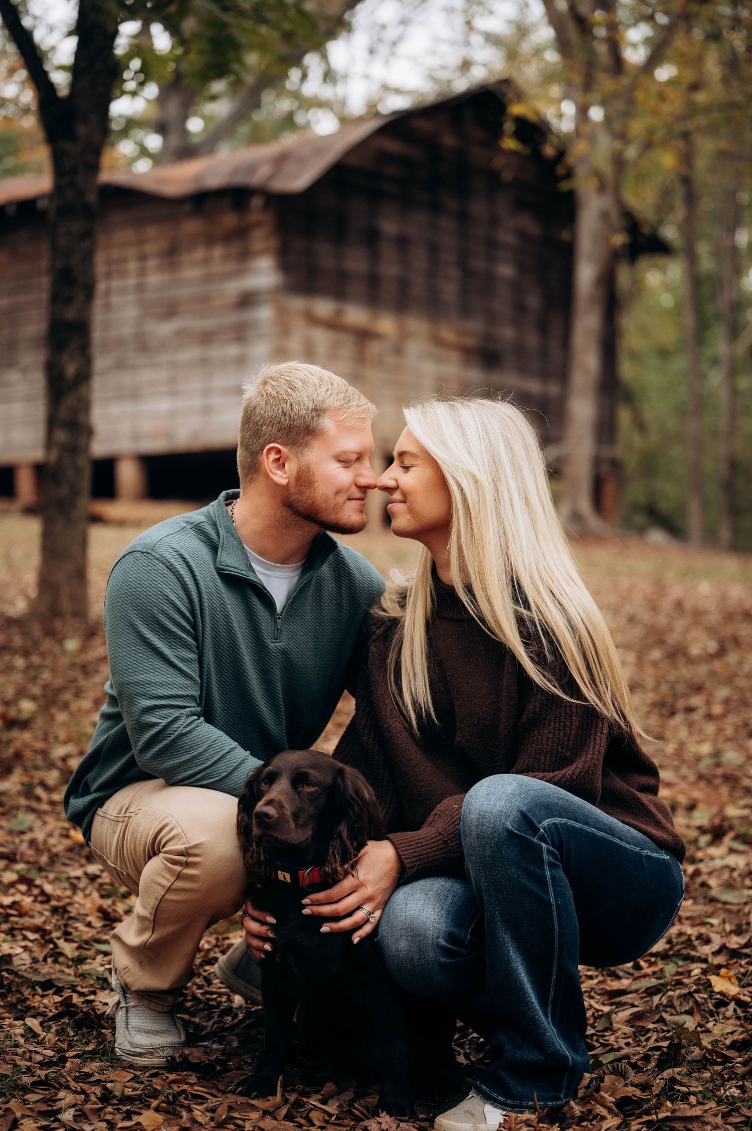 Couple sits with their black dog on fallen leaves near rustic wooden barn.