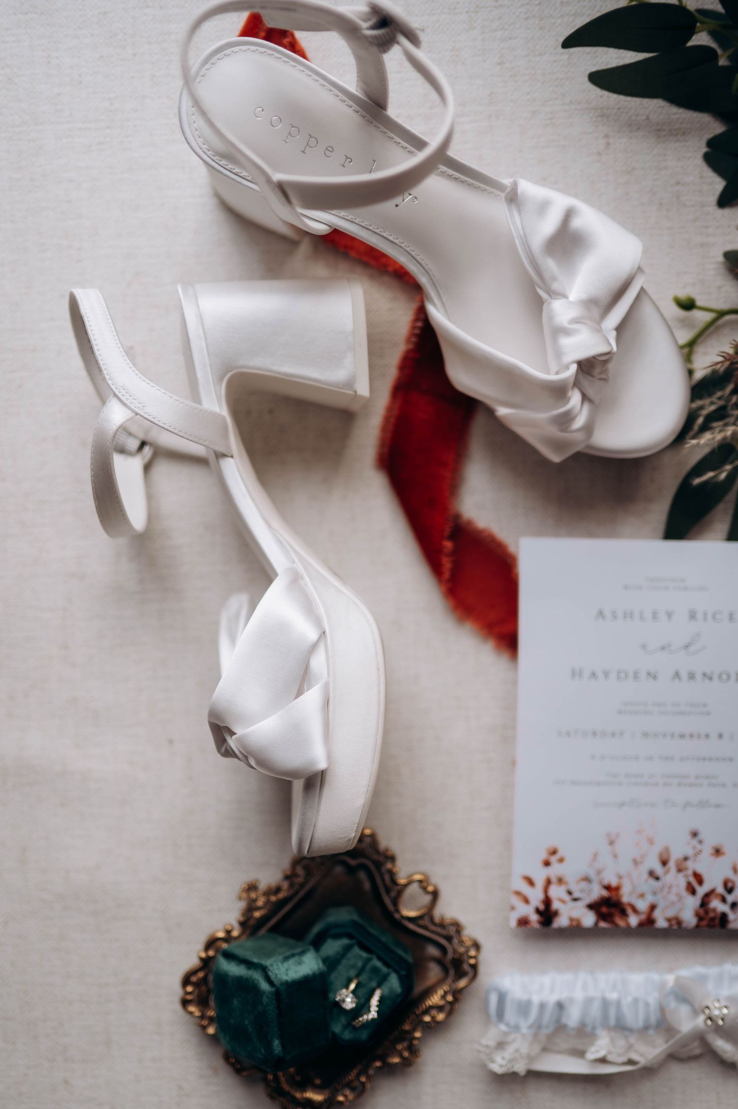 Wedding accessories including white shoes and stationery arranged with emerald velvet ring box.