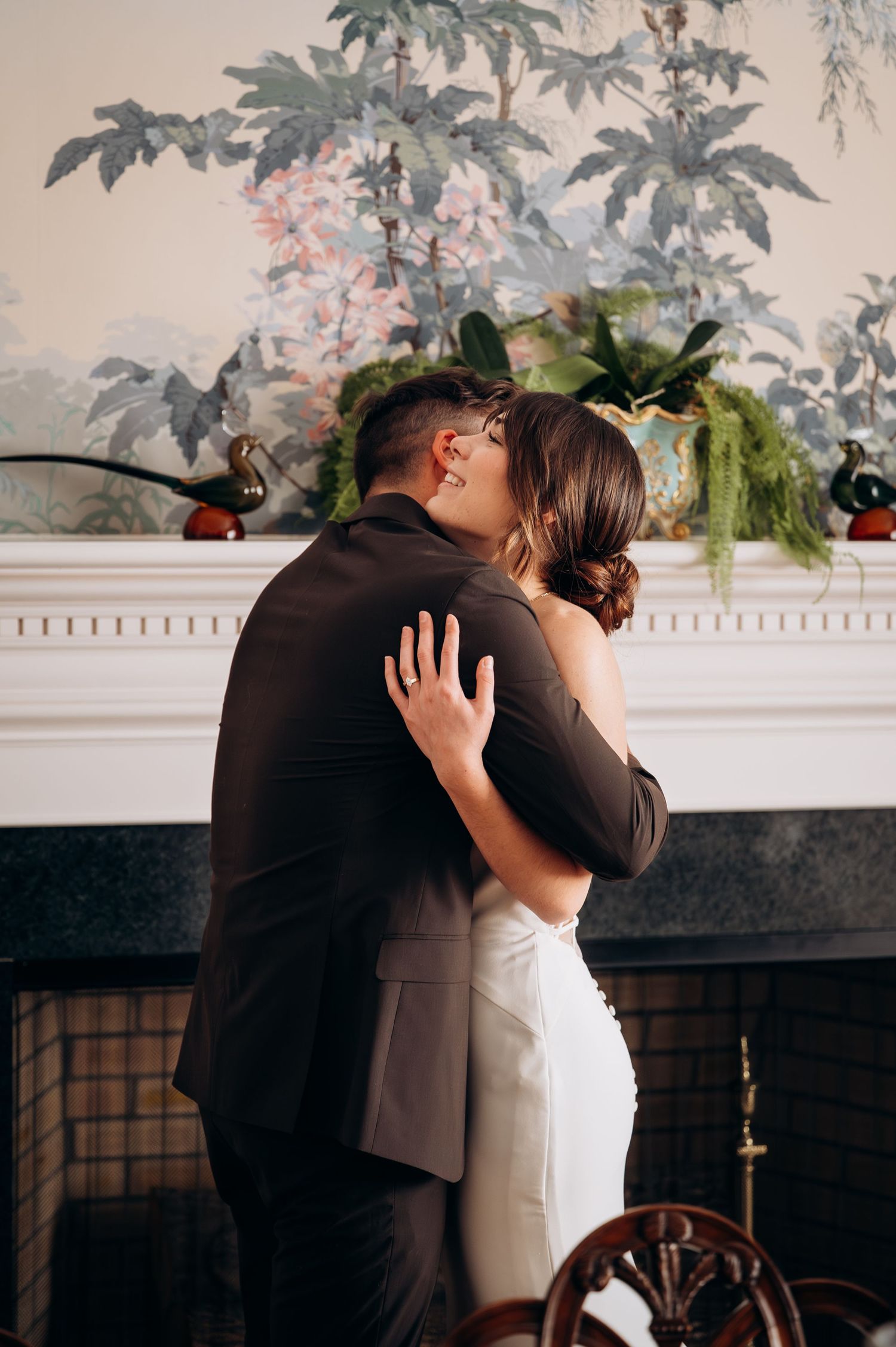 Couple embraces in an intimate moment in front of an elegant fireplace with floral wallpaper.
