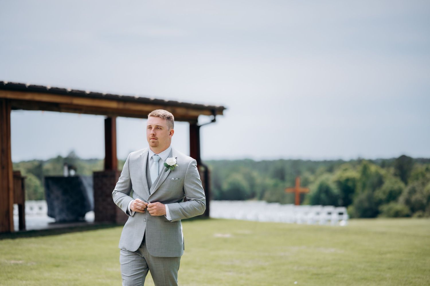 Groom in a light gray suit standing outdoors at a wedding venue with white chairs and a pavilion in the background.