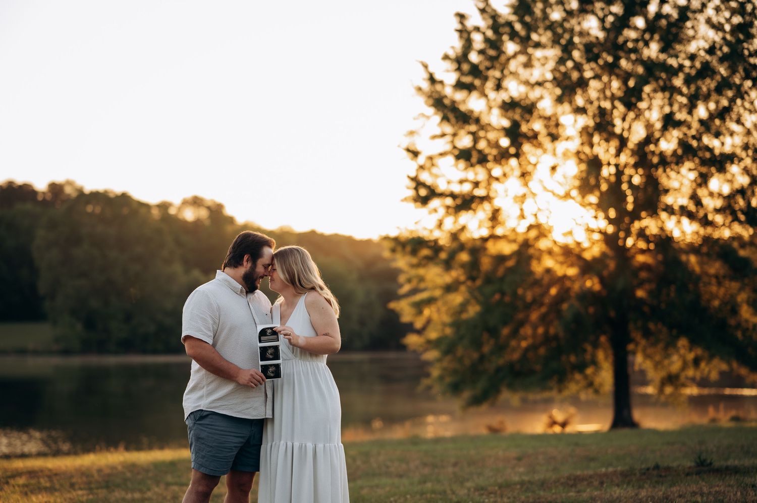Couple sharing a tender moment at golden hour by a lake, woman in white dress holding ultrasound photo.