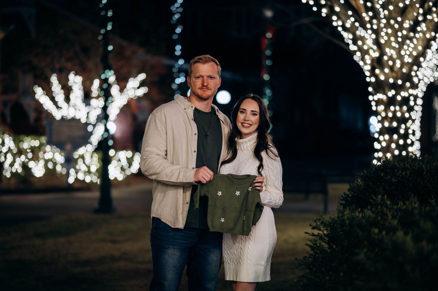 Couple posing together outdoors at night surrounded by festive holiday lights.